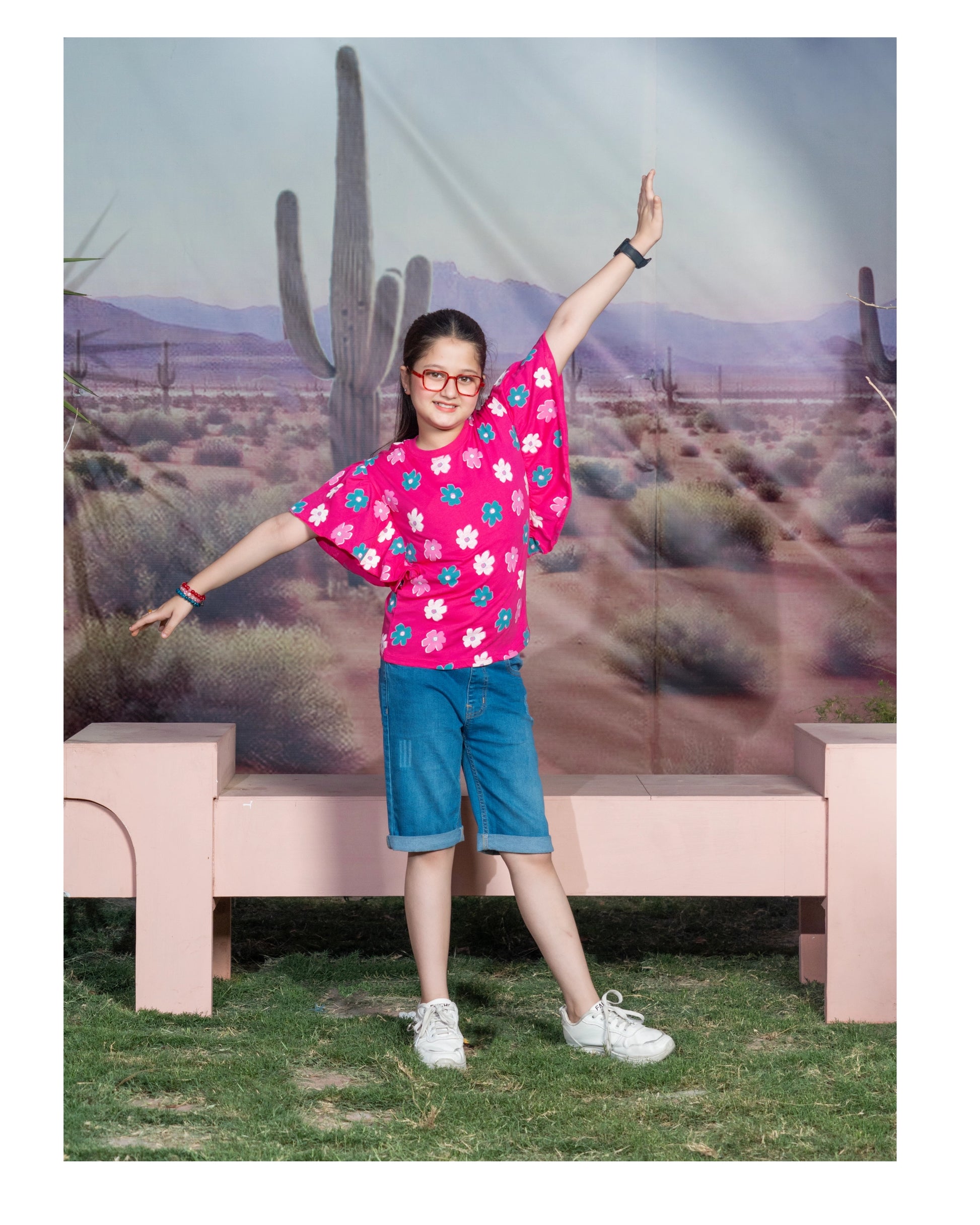 A young girl posing in a desert-themed studio setting, wearing a bright pink short-sleeve t-shirt with a colorful floral pattern, blue denim shorts, and white sneakers.