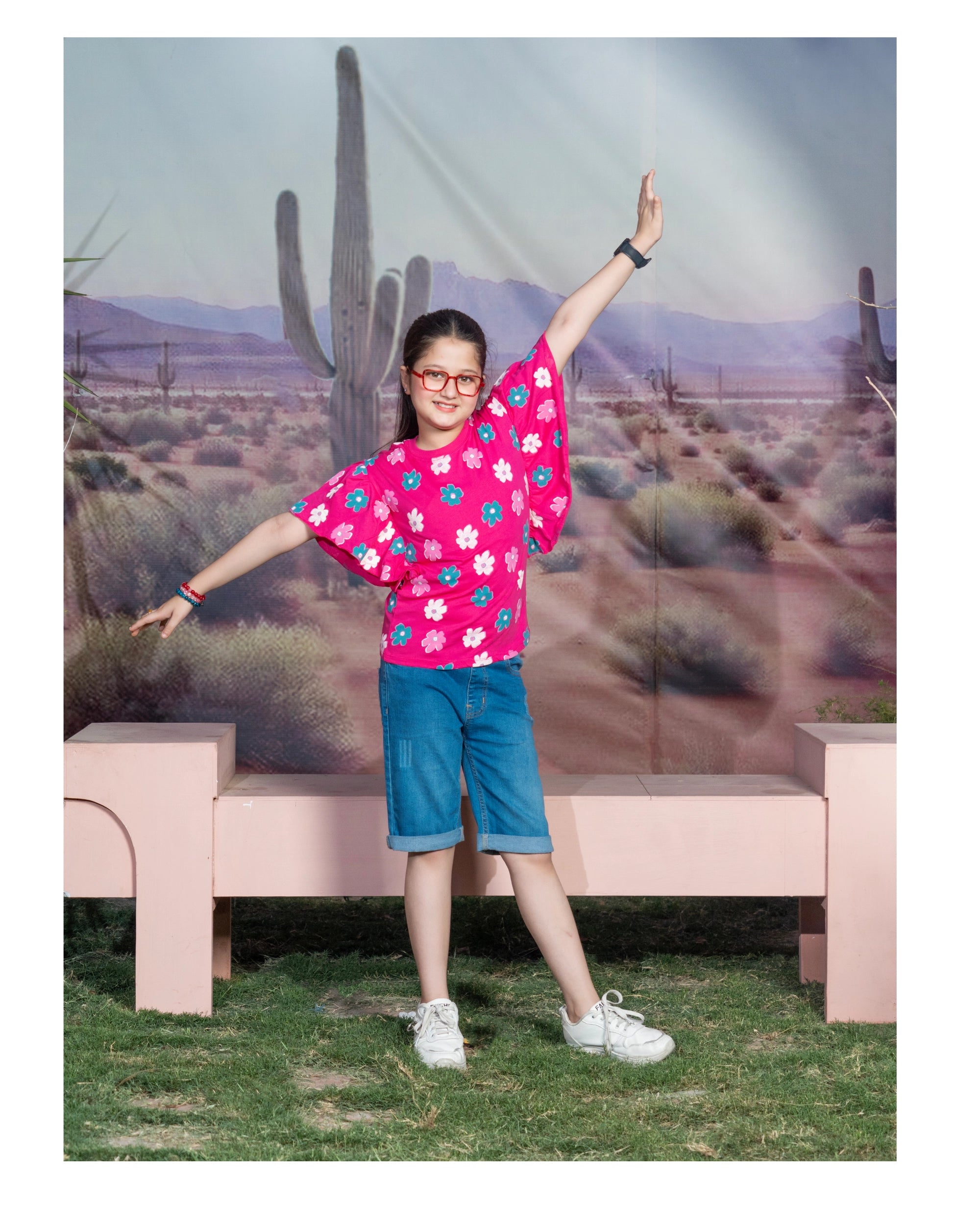 A young girl posing in a desert-themed studio setting, wearing a bright pink short-sleeve t-shirt with a colorful floral pattern, blue denim shorts, and white sneakers.