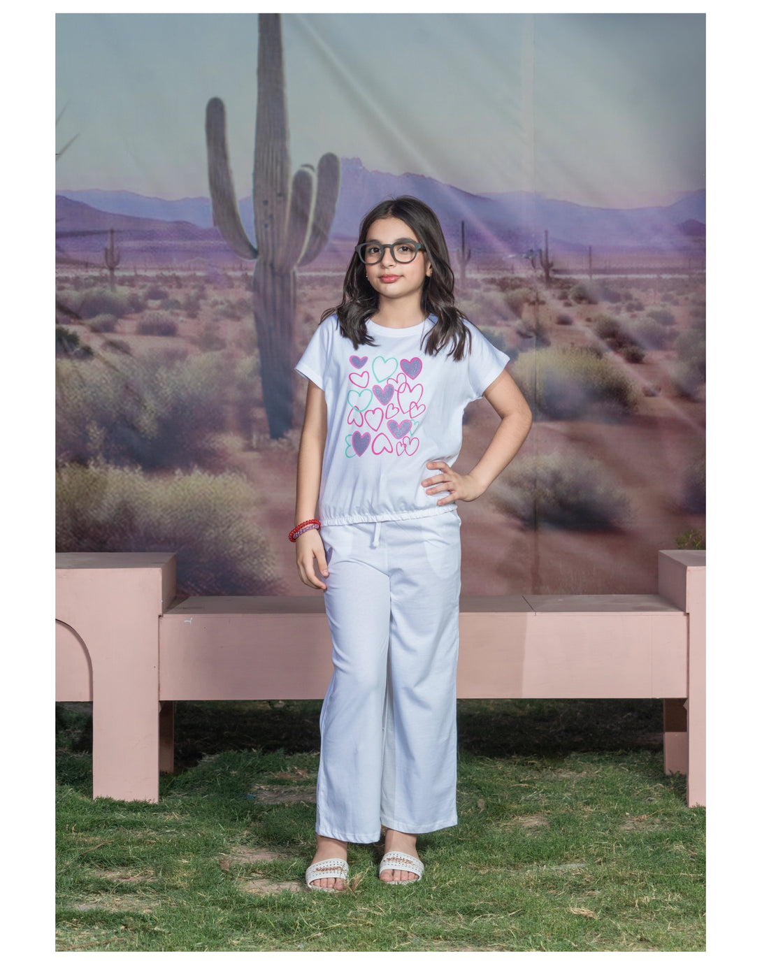Young girl wearing glasses and a light blue pajama set with a pink and purple “LOVE” heart graphic T-shirt and matching wide-leg pants, standing in front of a desert backdrop.