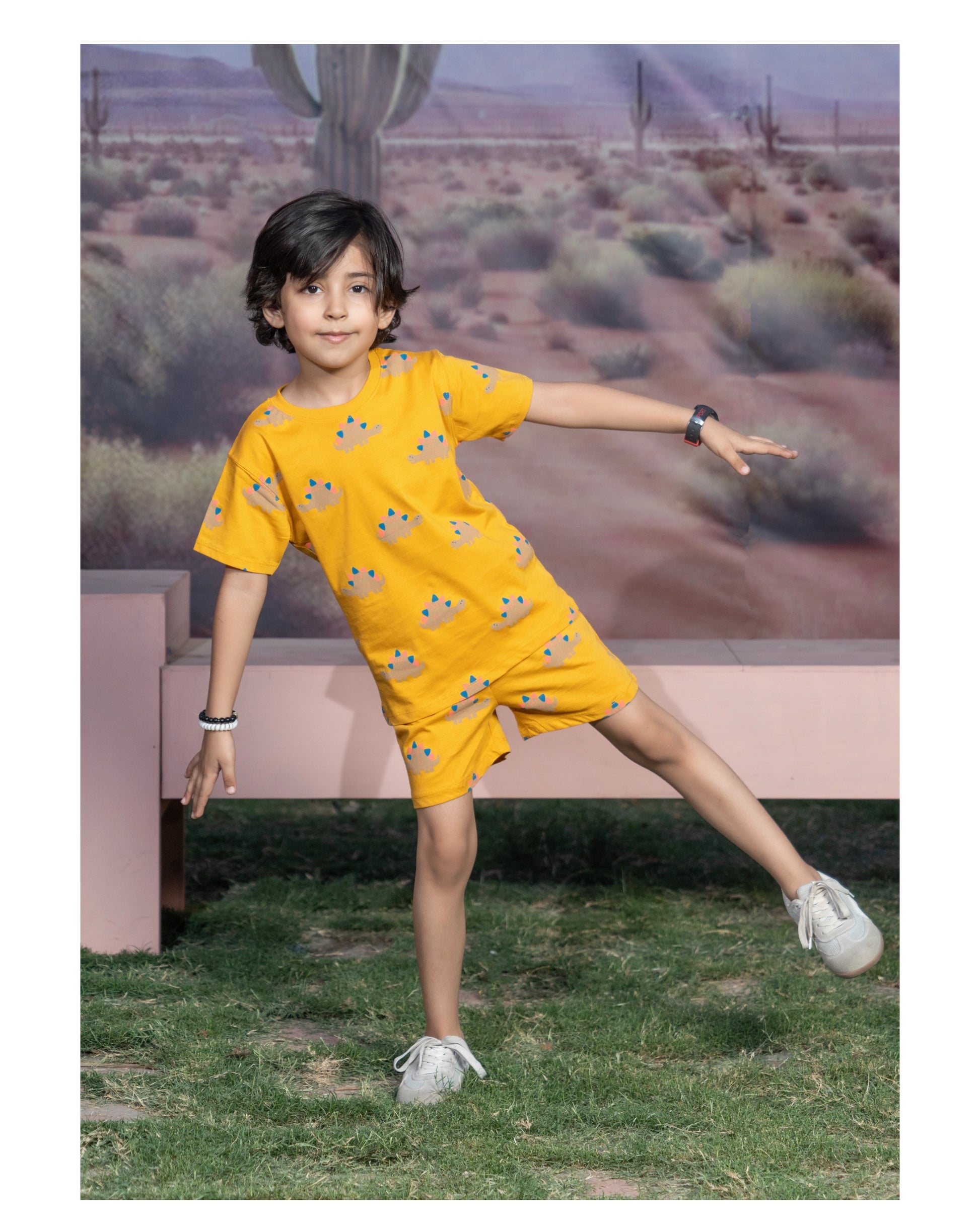 A young boy modeling a bright yellow short-sleeve tunic and shorts set with a cartoon animal print, posed in front of a desert landscape backdrop.
