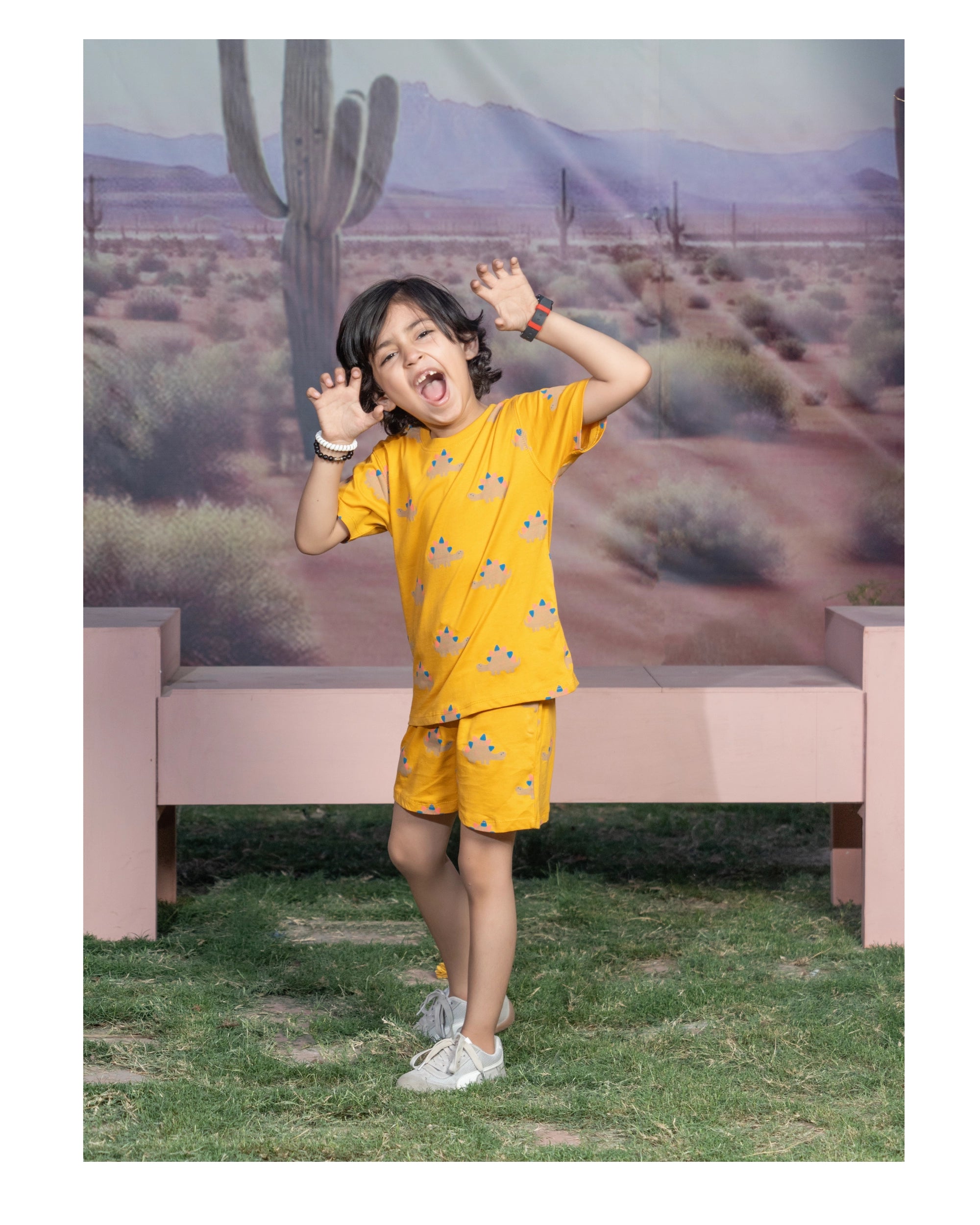 A young boy wearing a yellow short-sleeve pajama set with a cartoon print, posing in front of a desert landscape backdrop with a pink bench.