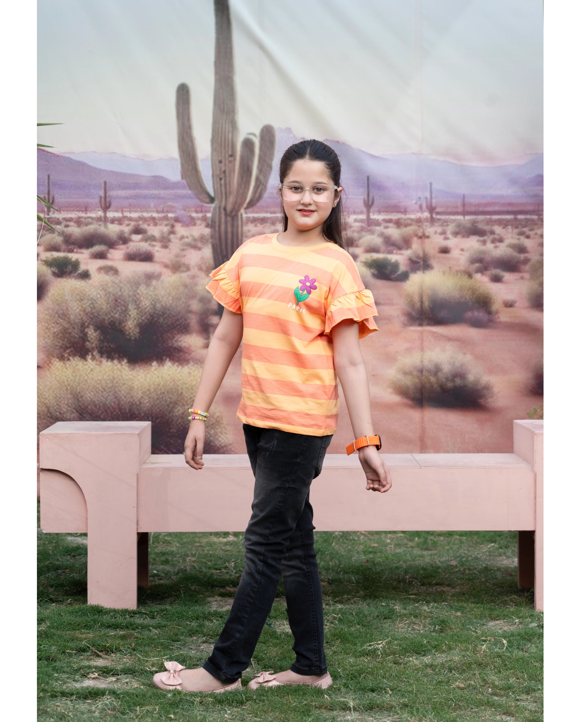 A young girl wearing a bright orange and yellow striped t-shirt with a small colorful graphic on the chest, paired with black pants, standing in front of a desert-themed backdrop.