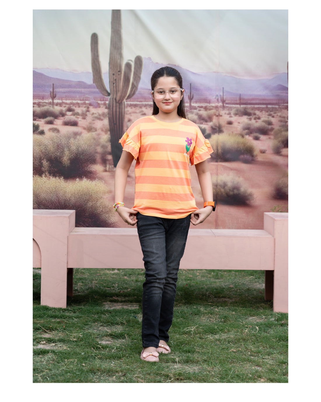 A young girl standing outdoors in front of a desert landscape backdrop, wearing a bright orange and yellow striped short-sleeve t-shirt with a small floral graphic on the chest, paired with dark jeans and pink sandals.