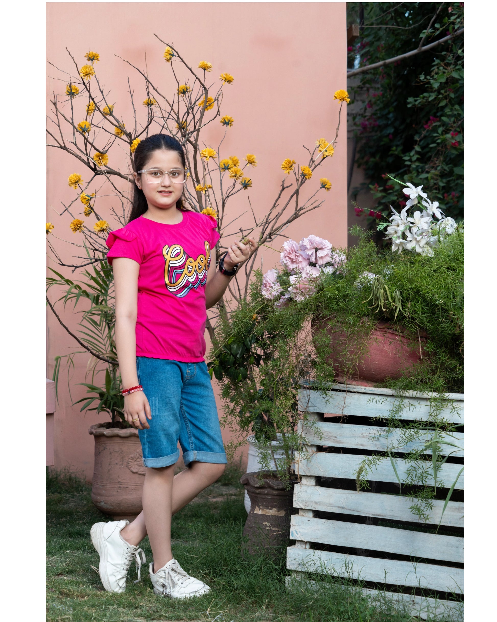 A young girl wearing a bright pink t-shirt with a colorful graphic and blue denim shorts, standing outdoors in front of a peach-colored wall with potted plants.