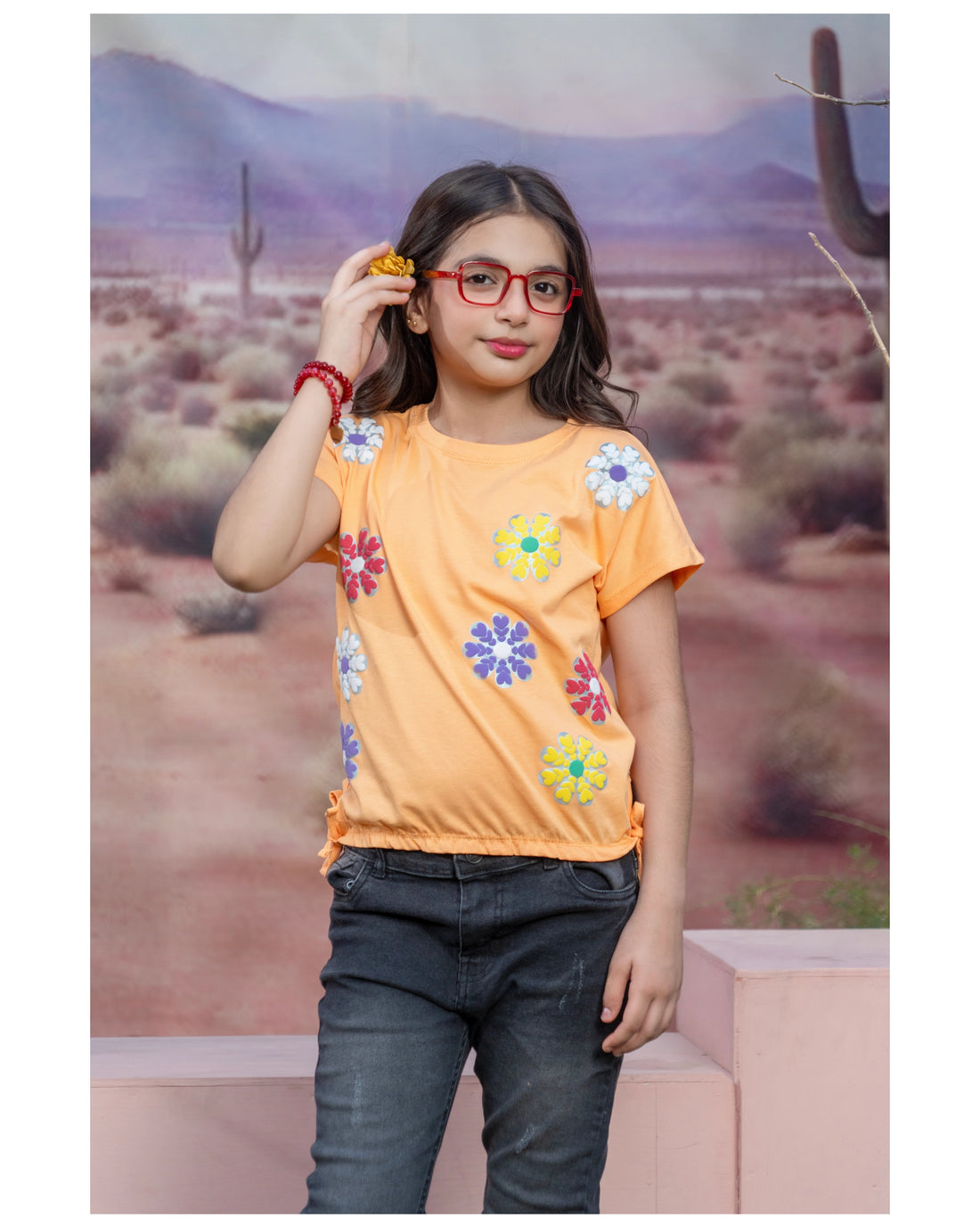 A young girl modeling a short-sleeve orange t-shirt with colorful embroidered flowers, paired with dark jeans, standing in front of a desert landscape backdrop.