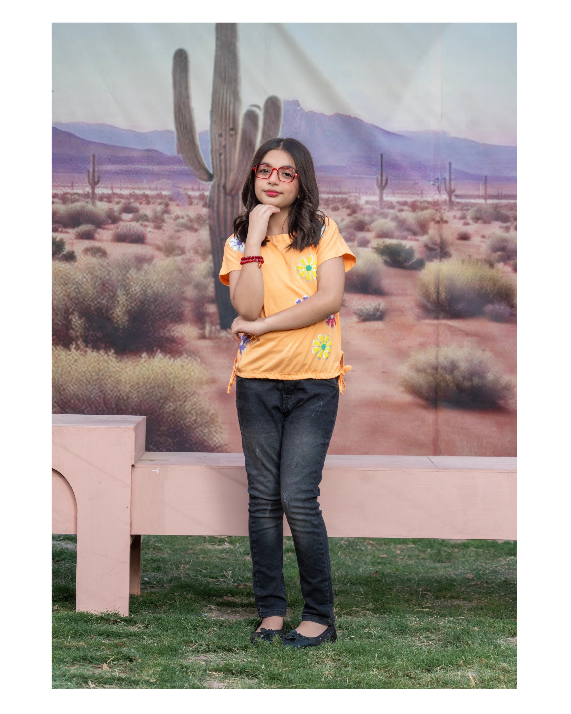 A woman wearing a bright orange short-sleeve t-shirt with colorful embroidered details, dark gray jeans, and black flats, posing in front of a desert landscape backdrop featuring saguaro cacti.